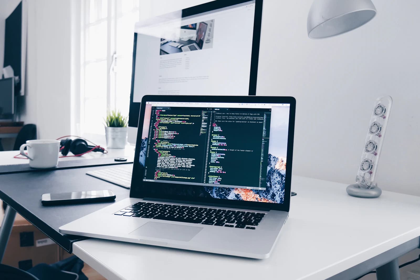 A laptop showing code on a bright desk beside a monitor, a coffee cup and headphones