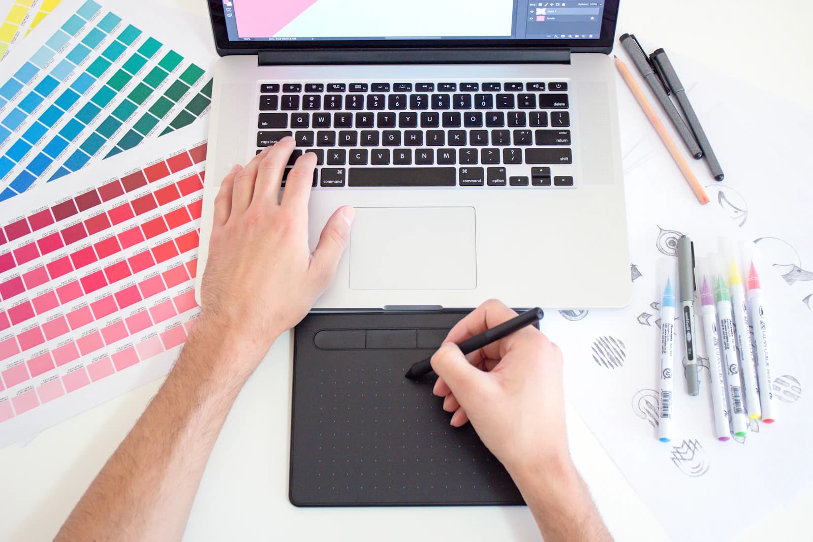 A designer drawing on a tablet beside a laptop, colour swatches and markers on the desk