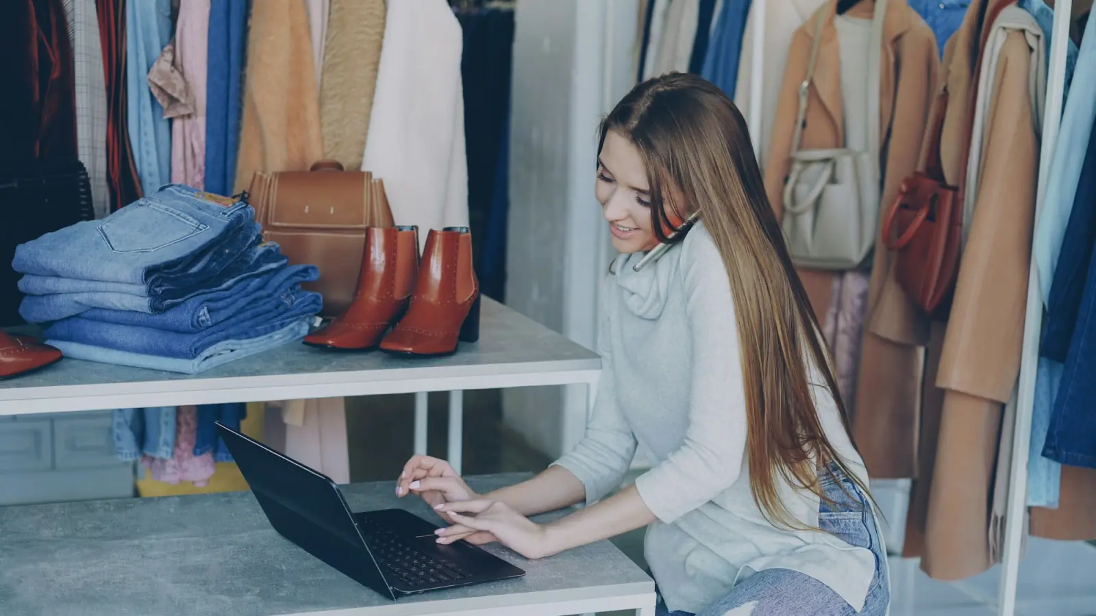 A boutique owner works on her laptop among racks of clothes, shoes and folded jeans in her shop.