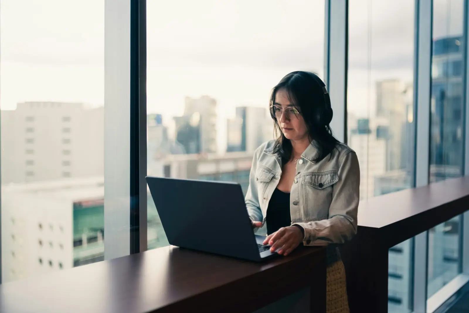 A woman working on her laptop at a desk by a window over a city skyline