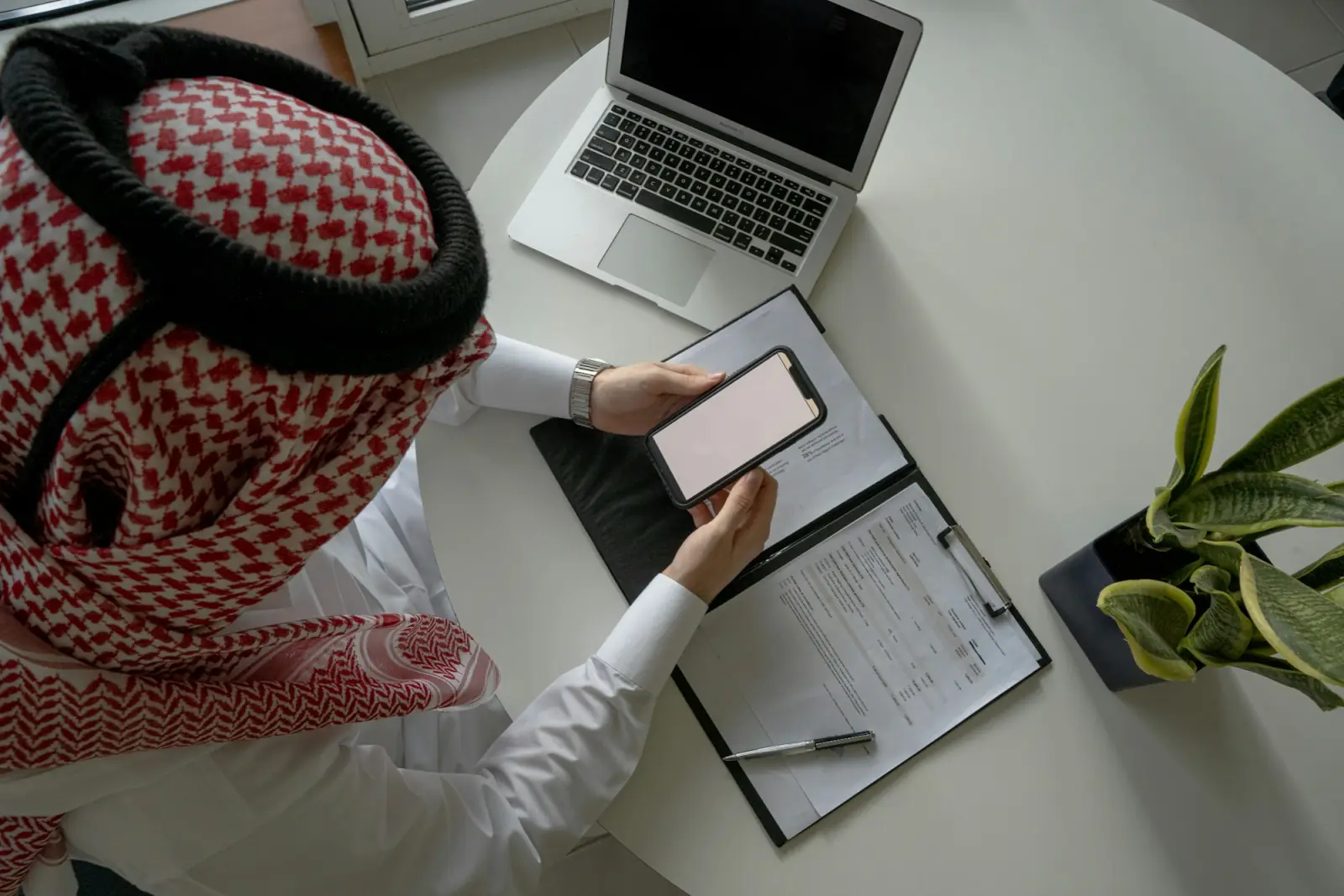 A business owner in a ghutra answers a customer message on his phone at his desk, laptop and notebook beside him