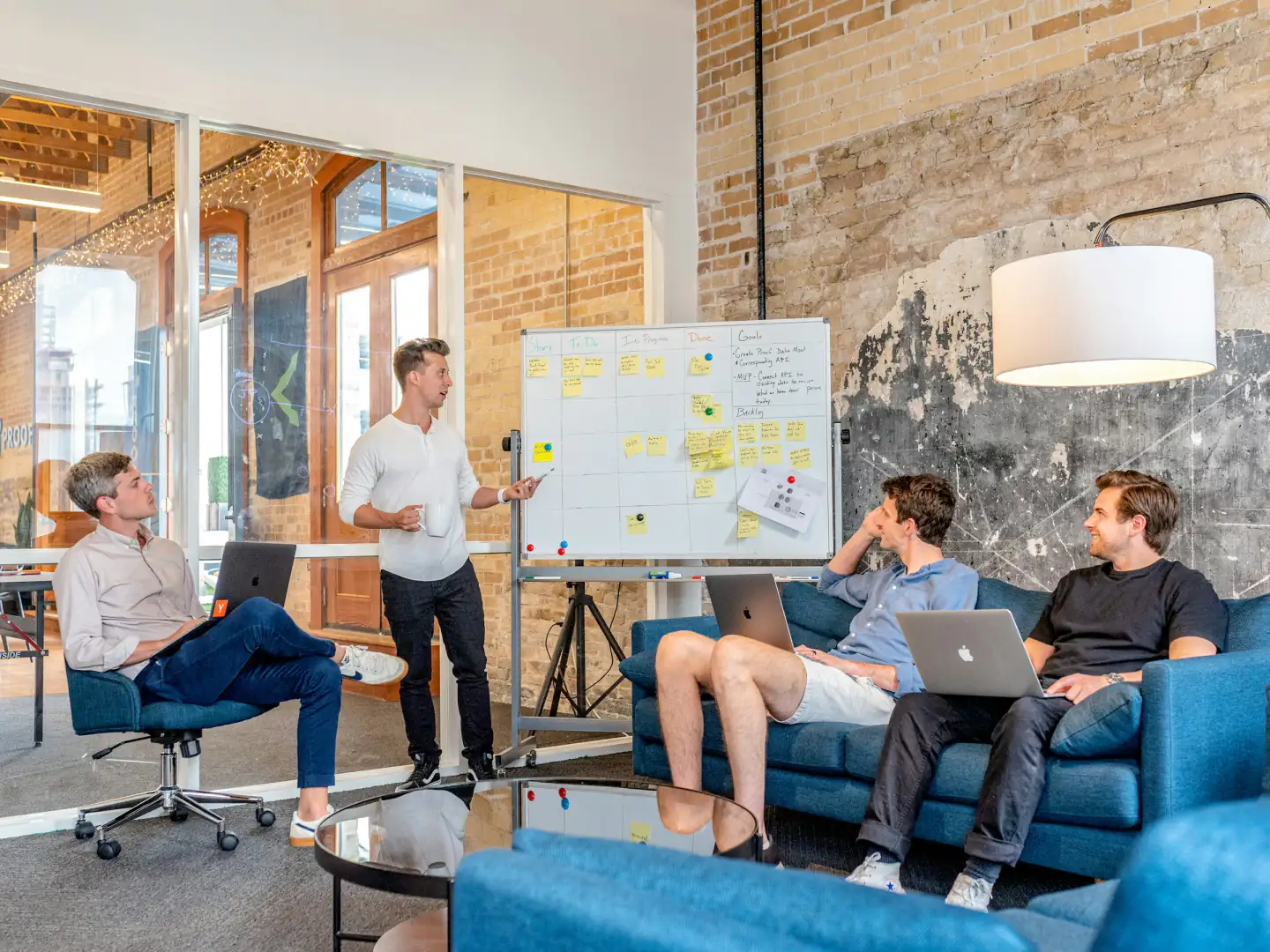 A team planning a project at a whiteboard covered in sticky notes, laptops open on the sofa