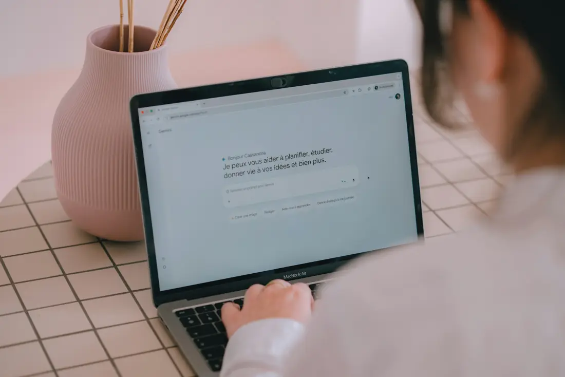 A woman typing into an assistant on her laptop at a light, tidy desk