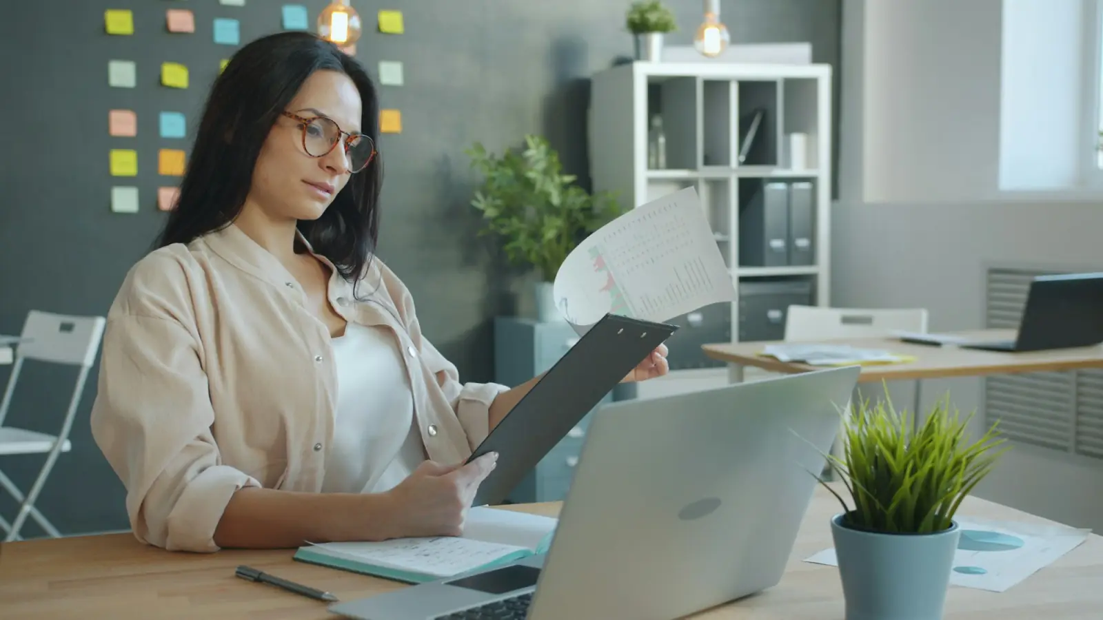 A woman at her desk reading through printed documents beside her open laptop