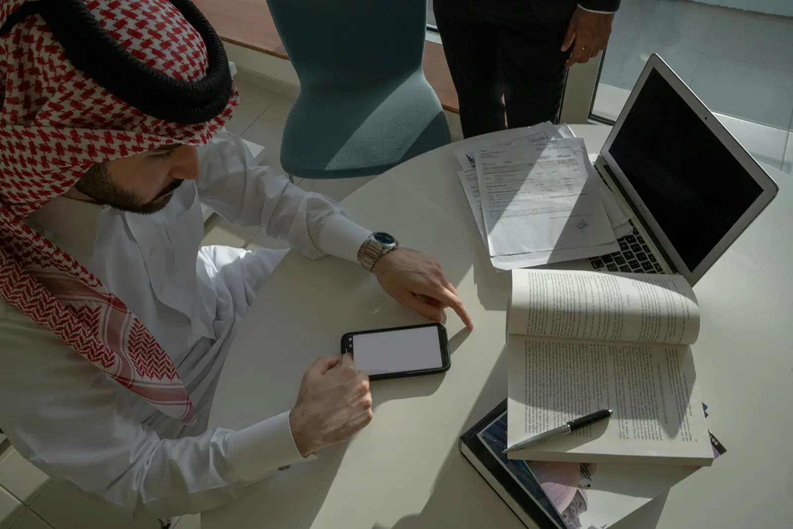 A business owner in a ghutra plans at his desk with his laptop, phone and notebook