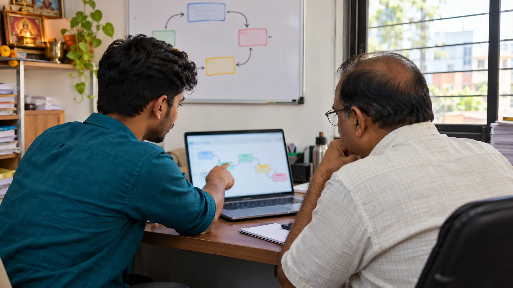 A young developer points at a chain of connected steps on a laptop while a business owner looks on, a loop of boxes and arrows drawn on the whiteboard behind them.