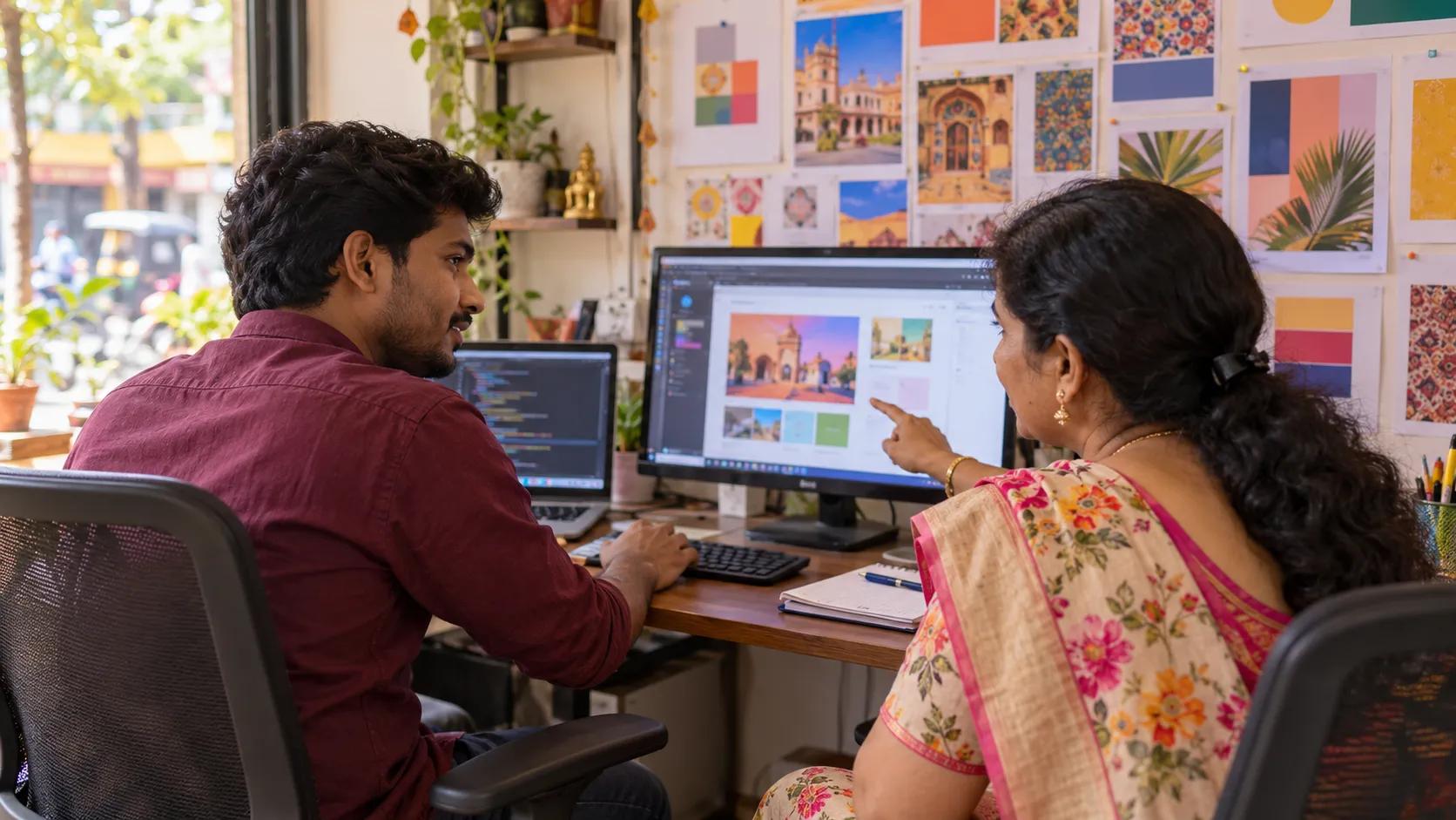 A shop owner in a floral saree points at a website editor on a monitor while a web developer, with code open on the laptop beside it, turns to answer.