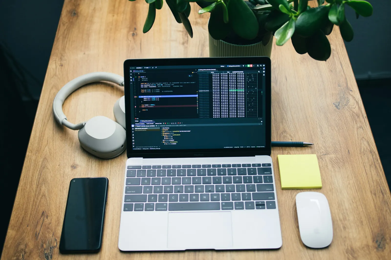 A laptop showing website code on a wooden desk, with headphones, a phone, a notebook and sticky notes beside it