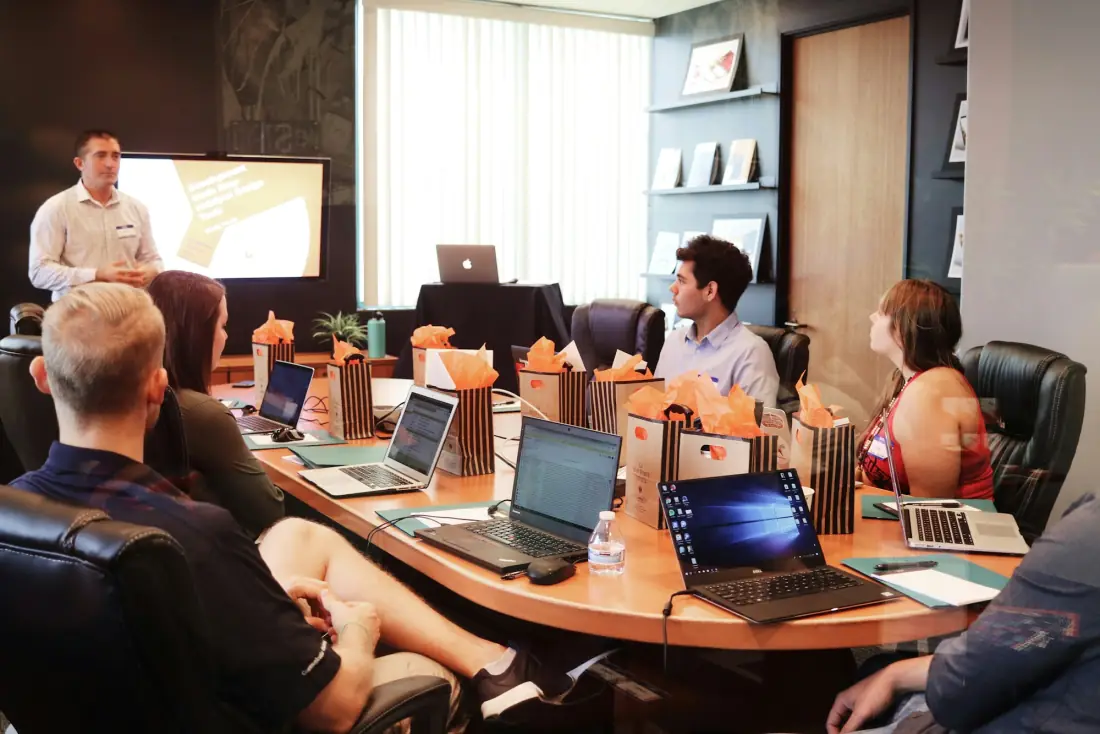A marketing team around a meeting table with laptops open while one person presents