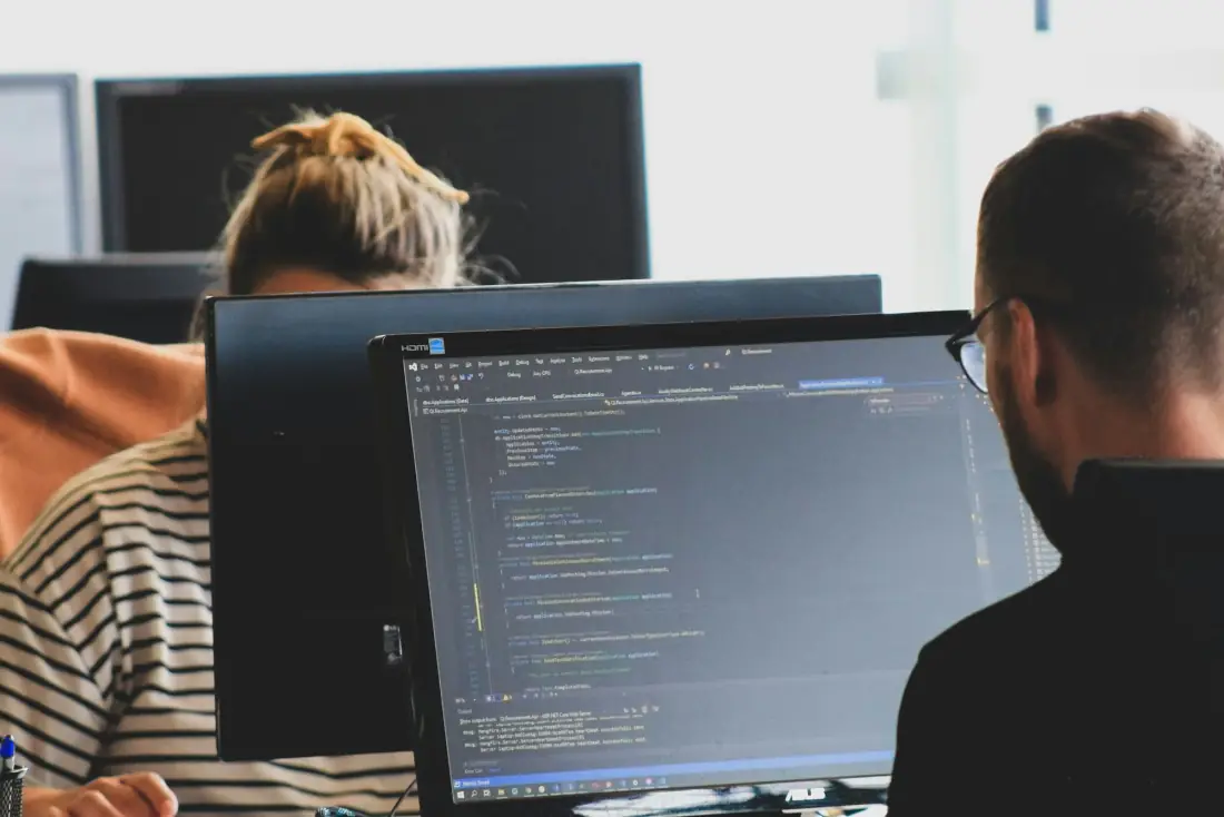 Two developers writing code side by side at their monitors in a bright office