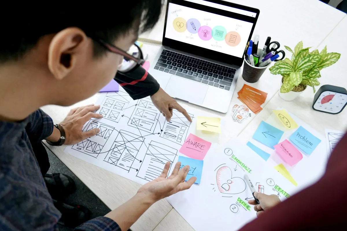A web designer sketching page wireframes on paper beside a laptop, with sticky notes and pens across the desk